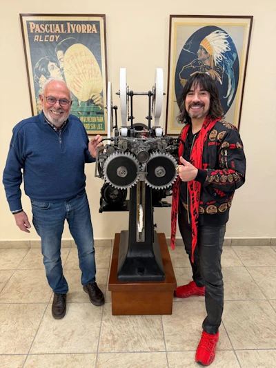 Two men pose with a vintage mechanical gear device in front of classic Spanish advertising posters