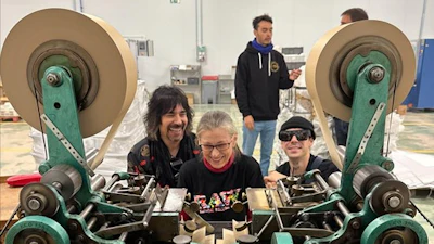 Three people pose in front of a large turquoise industrial printing press machine in a workshop facility