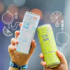 Two hands holding CANN natural tonic cans in blue grapefruit rosemary and yellow lemon lavender flavors at an outdoor event