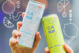 Two hands holding CANN natural tonic cans in blue grapefruit rosemary and yellow lemon lavender flavors at an outdoor event