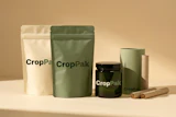 CropPak sustainable packaging collection featuring cream and green pouches, black glass jar, and cardboard tube on neutral background