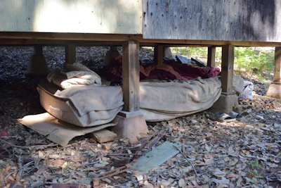 Makeshift shelter with bedding and belongings under a wooden structure in outdoor setting