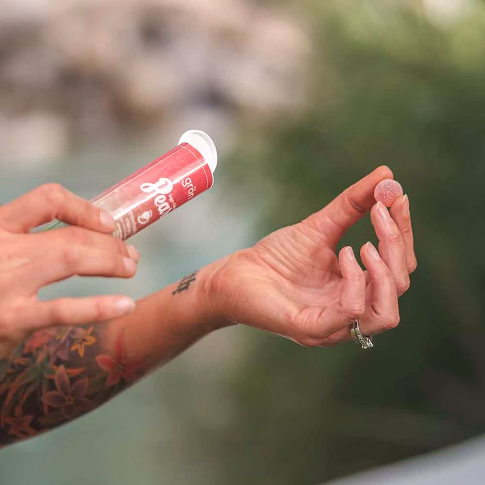Person applying pink lip gloss with visible tattoo on arm against blurred outdoor background