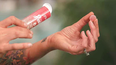 Person applying pink lip gloss with visible tattoo on arm against blurred outdoor background