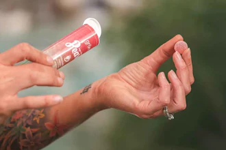 Person applying pink lip gloss with visible tattoo on arm against blurred outdoor background