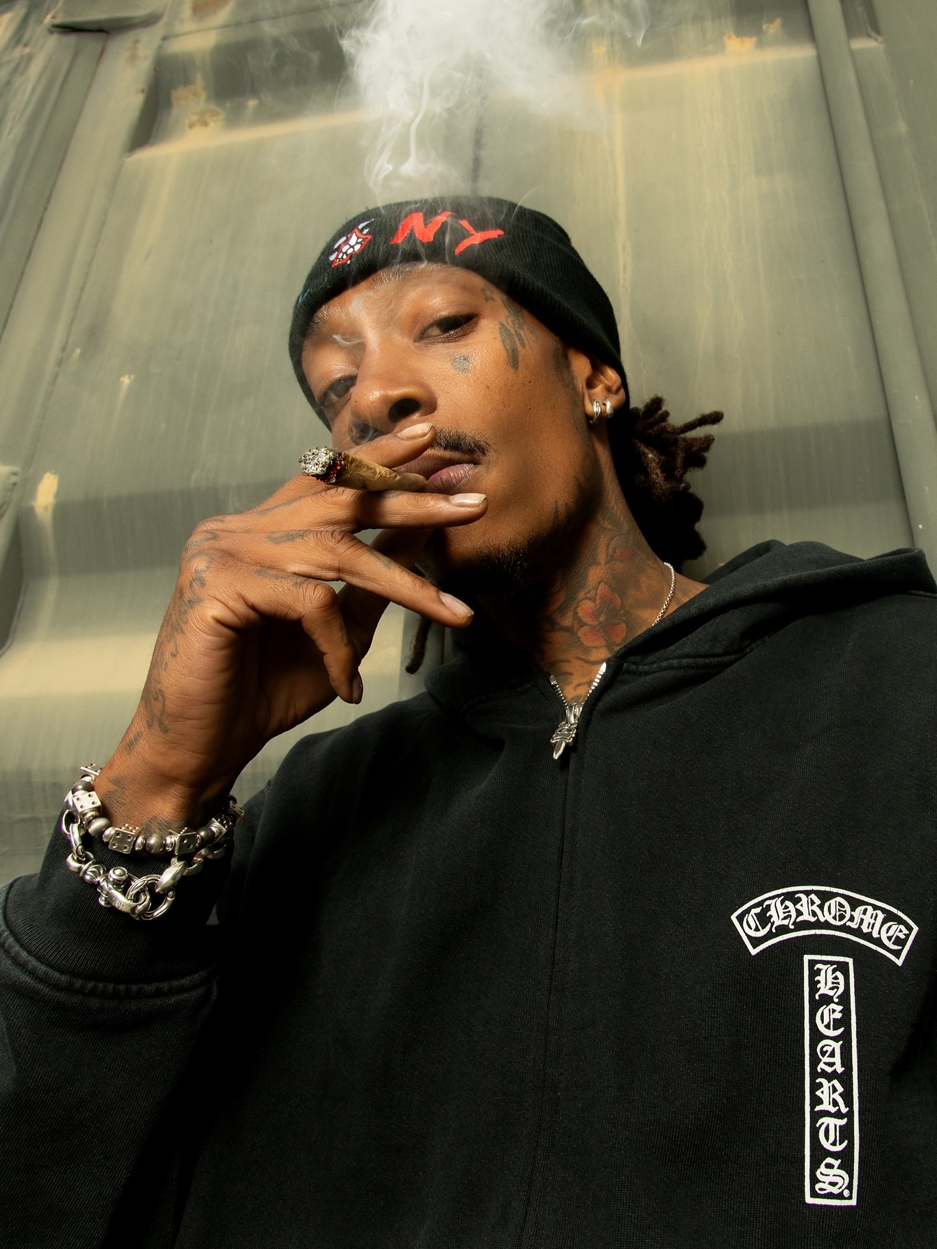 Person wearing black Carhartt hoodie smoking a cigar, with face tattoos and gold jewelry, posed indoors