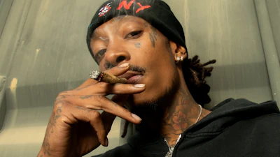 Person wearing black Carhartt hoodie smoking a cigar, with face tattoos and gold jewelry, posed indoors