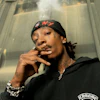 Person wearing black Carhartt hoodie smoking a cigar, with face tattoos and gold jewelry, posed indoors