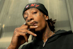Person wearing black Carhartt hoodie smoking a cigar, with face tattoos and gold jewelry, posed indoors