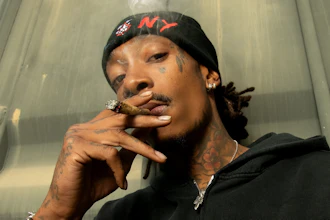Person wearing black Carhartt hoodie smoking a cigar, with face tattoos and gold jewelry, posed indoors