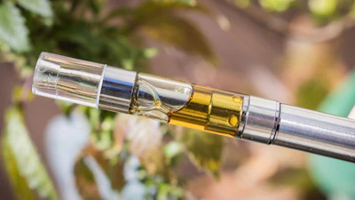 Golden vaping cartridge attached to silver battery component with blurred green foliage background