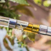 Golden vaping cartridge attached to silver battery component with blurred green foliage background