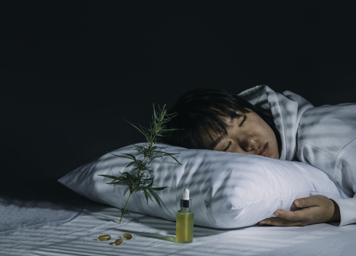 Person resting head on white pillow with cannabis plant and CBD oil bottle on bed against dark background