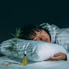 Person resting head on white pillow with cannabis plant and CBD oil bottle on bed against dark background