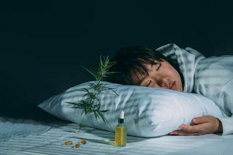 Person resting head on white pillow with cannabis plant and CBD oil bottle on bed against dark background