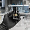Black cosmetic bottle with gold leaf logo held in gloved hand in laboratory setting