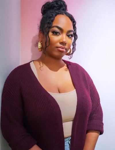 Woman wearing burgundy cardigan and gold hoop earrings poses against pink and white gradient background