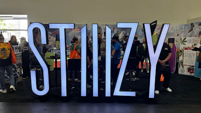 Large blue STIIIZY text installation at indoor event with attendees and branded merchandise displays