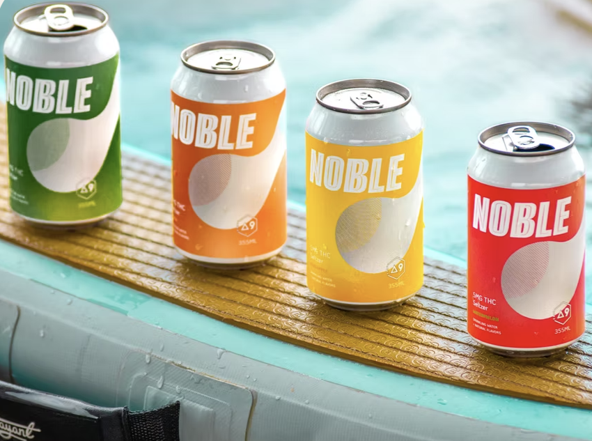 Four colorful Noble beverage cans in green, orange, yellow, and red arranged on a wooden surface near water