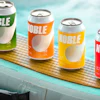 Four colorful Noble beverage cans in green, orange, yellow, and red arranged on a wooden surface near water