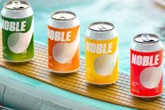 Four colorful Noble beverage cans in green, orange, yellow, and red arranged on a wooden surface near water